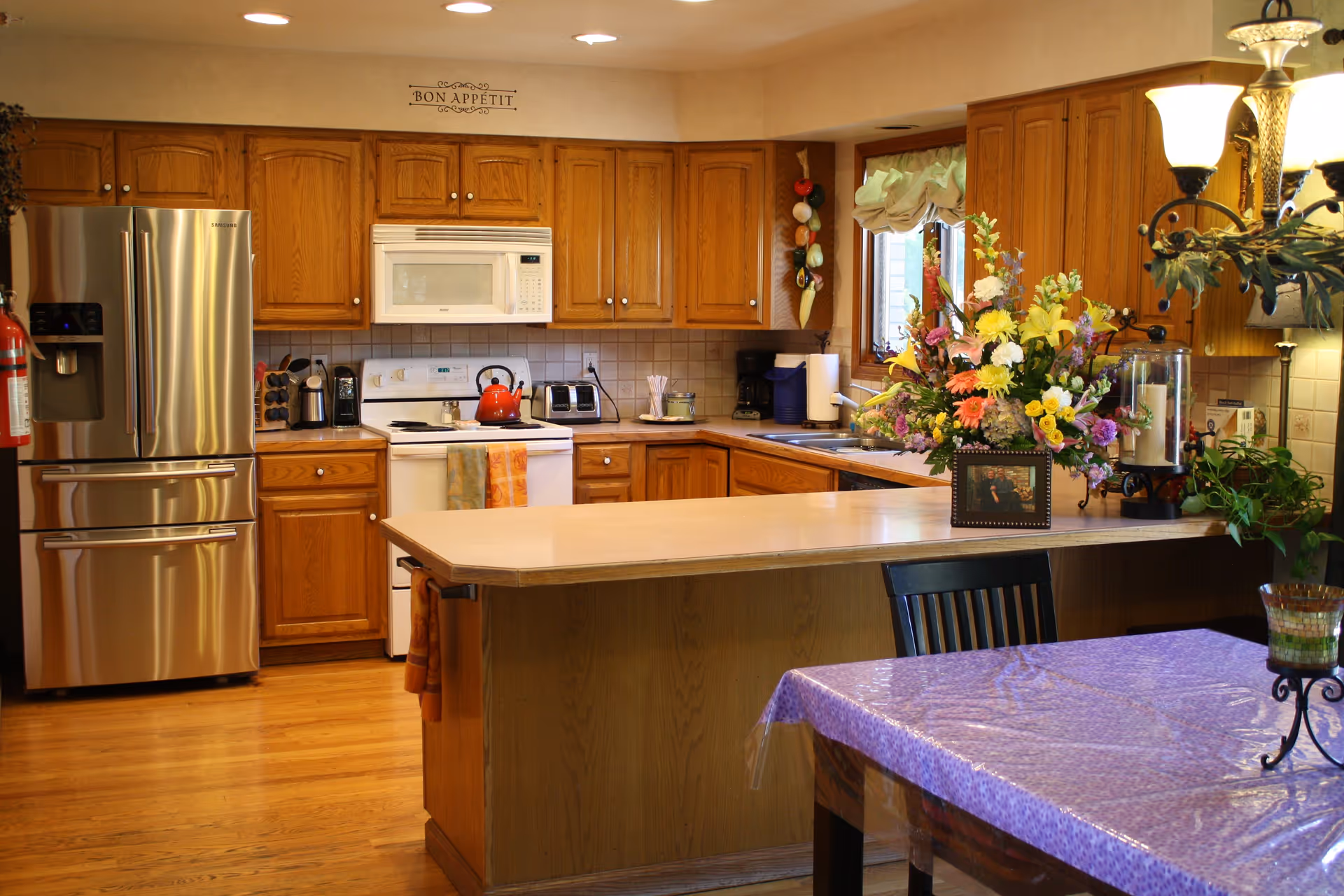 A warm and inviting kitchen with wooden cabinets, a stainless steel refrigerator, a white stove with a red kettle on it, and a white microwave above the stove. There is a kitchen island with a light-colored countertop and a bouquet of colorful flowers in a vase on it. A dining table with a purple tablecloth and a candle holder is partially visible in the foreground. The kitchen has wooden flooring and a window with a curtain letting in natural light. A decorative sign above the cabinets reads 'Bon Appétit'.