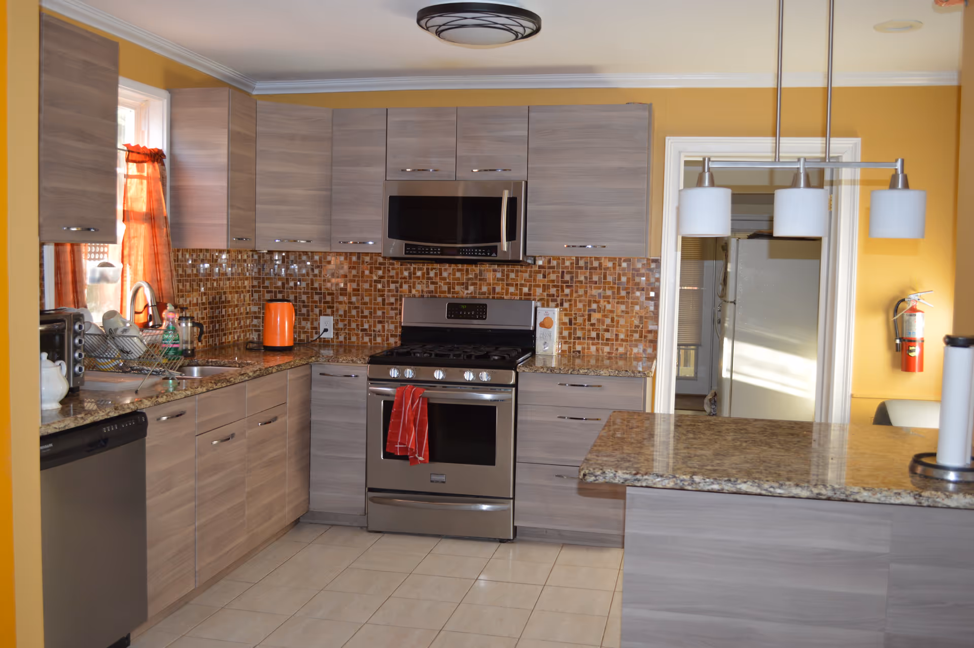 Modern kitchen with light wood cabinets, granite countertops, and a mosaic tile backsplash. Stainless steel appliances include a stove with a red towel hanging on the handle, a microwave above it, and a dishwasher. There is a window with orange curtains, a dish rack with dishes, an orange electric kettle, and a paper towel holder on the counter. The walls are painted yellow, and there is a three-light pendant fixture hanging above the counter.