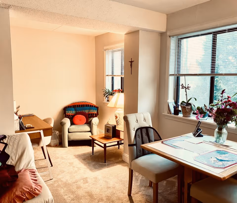 A cozy living room area in a retirement community featuring a beige armchair with a colorful blanket and red cushion, a wooden side table with a lamp, a desk with a chair, and a dining table with two upholstered chairs. The room has large windows with blinds, potted plants on the windowsill, and a crucifix on the wall.