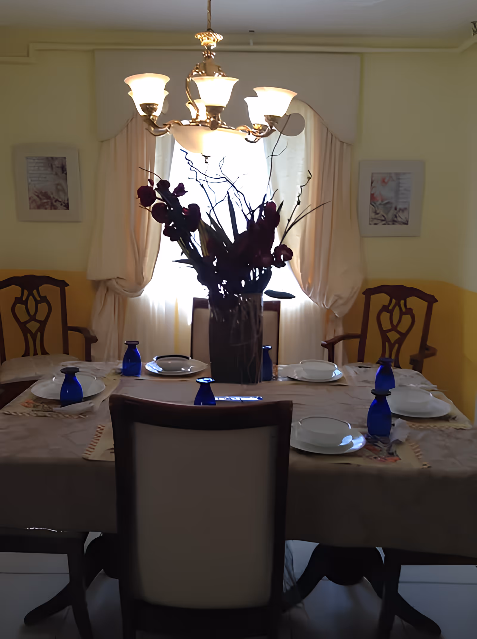 A dining room with a table set for six people, featuring white plates and blue glasses. There is a large floral centerpiece on the table. The room has yellow walls with two framed pictures and a window with cream-colored curtains. A chandelier with five lights hangs above the table.