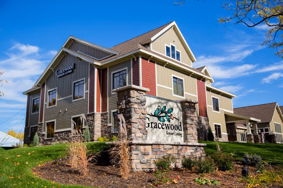 Exterior view of a two-story senior living facility building with beige and red siding, stone accents, and multiple windows under a clear blue sky. In front of the building is a stone sign with the Gracewood Senior Living logo featuring oak leaves and acorns.