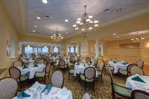 A spacious dining room with multiple round tables covered with white tablecloths, each set with green napkins, plates, cups, and silverware. The room features elegant chandeliers hanging from a white ceiling, beige walls, and large windows allowing natural light to enter.