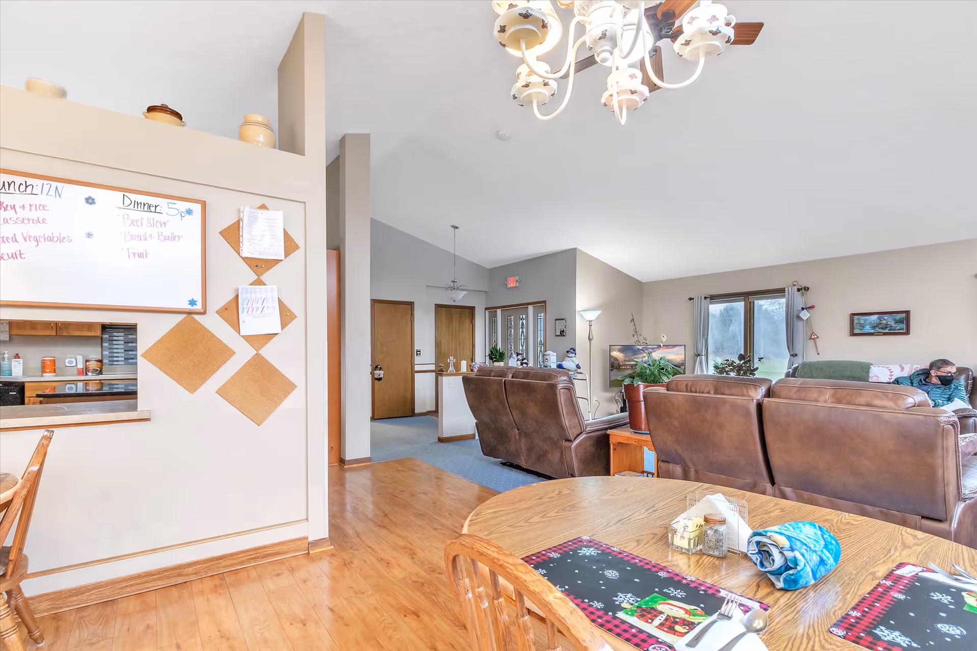 Interior view of a senior living facility showing a dining area with a wooden table set with placemats and utensils in the foreground, and a living room with brown leather sofas, a TV, and a person sitting on a couch in the background. A whiteboard with lunch and dinner menus is mounted on a wall near the dining area. The room has wooden flooring and neutral-colored walls with a chandelier hanging from the ceiling.