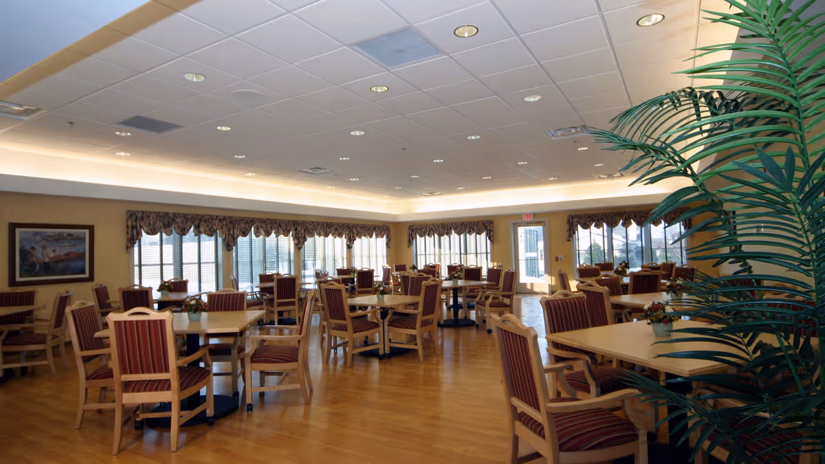 Spacious dining room with multiple wooden tables and striped upholstered chairs, large windows, and a potted plant.