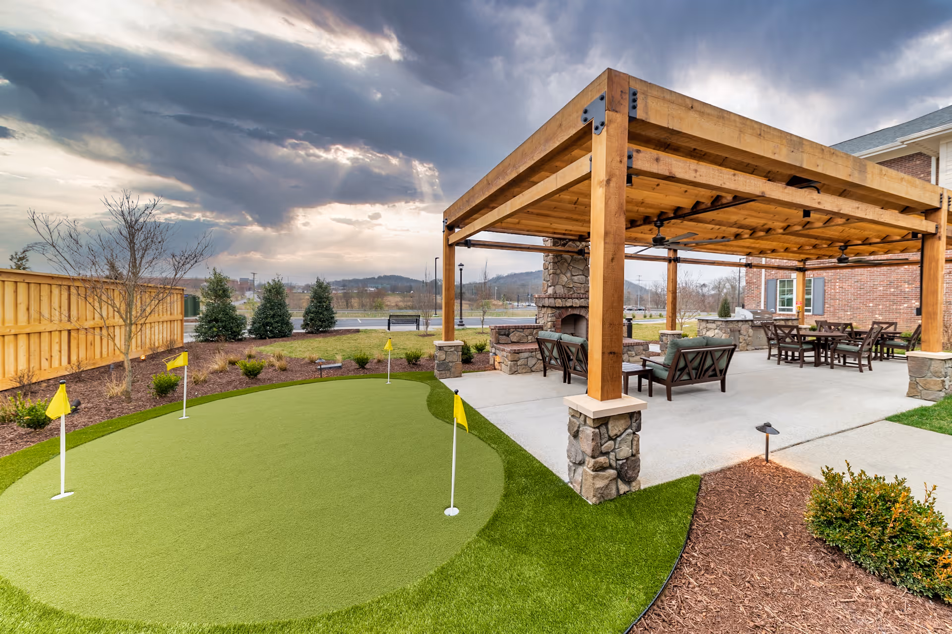 Outdoor patio area at The Capstone at Station Camp featuring a wooden pergola with stone pillars, outdoor seating with cushioned chairs and tables, a stone fireplace, and a small putting green with four yellow flags. The area is surrounded by landscaping with bushes, a wooden fence, and a cloudy sky overhead.
