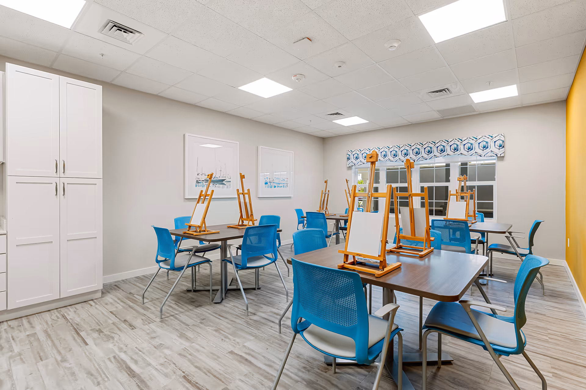 A bright, clean art activity room with multiple tables and blue chairs. Each table has wooden easels with blank canvases set up for painting. The room has light-colored walls, a yellow accent wall, framed artwork, and a window with a patterned valance.