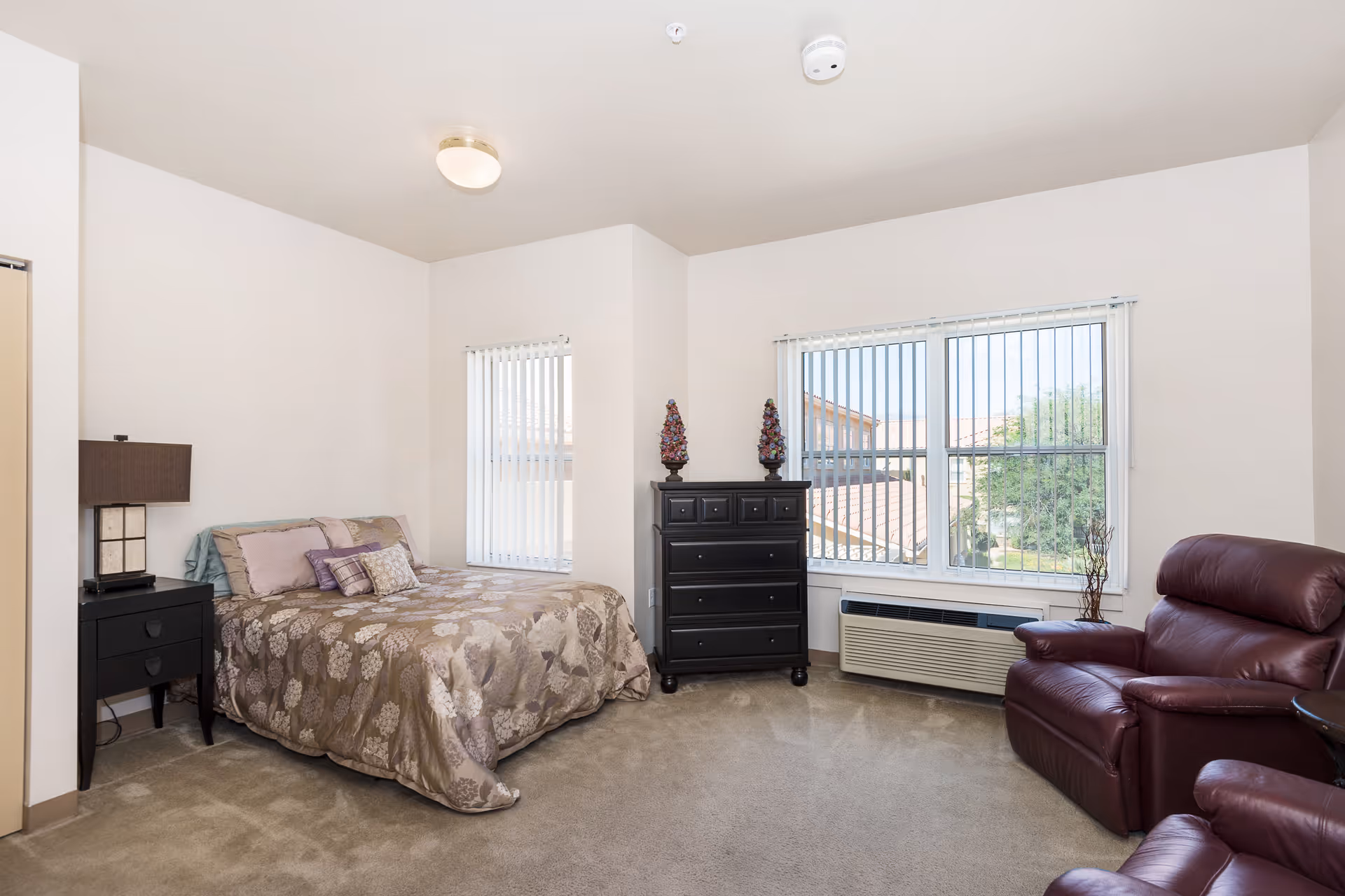 A bright bedroom with a bed covered in a floral patterned comforter, a black nightstand with a lamp, a black chest of drawers with decorative items on top, a large window with vertical blinds, and a burgundy leather recliner chair.