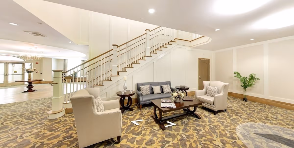 A spacious and well-lit common area with a staircase in the background. The seating area includes a gray sofa and two beige armchairs arranged around a wooden coffee table with magazines and a floral centerpiece. There are two side tables with lamps, a potted plant in the corner, and patterned carpet flooring. The entrance area with glass doors and a round table with flowers is visible in the background.