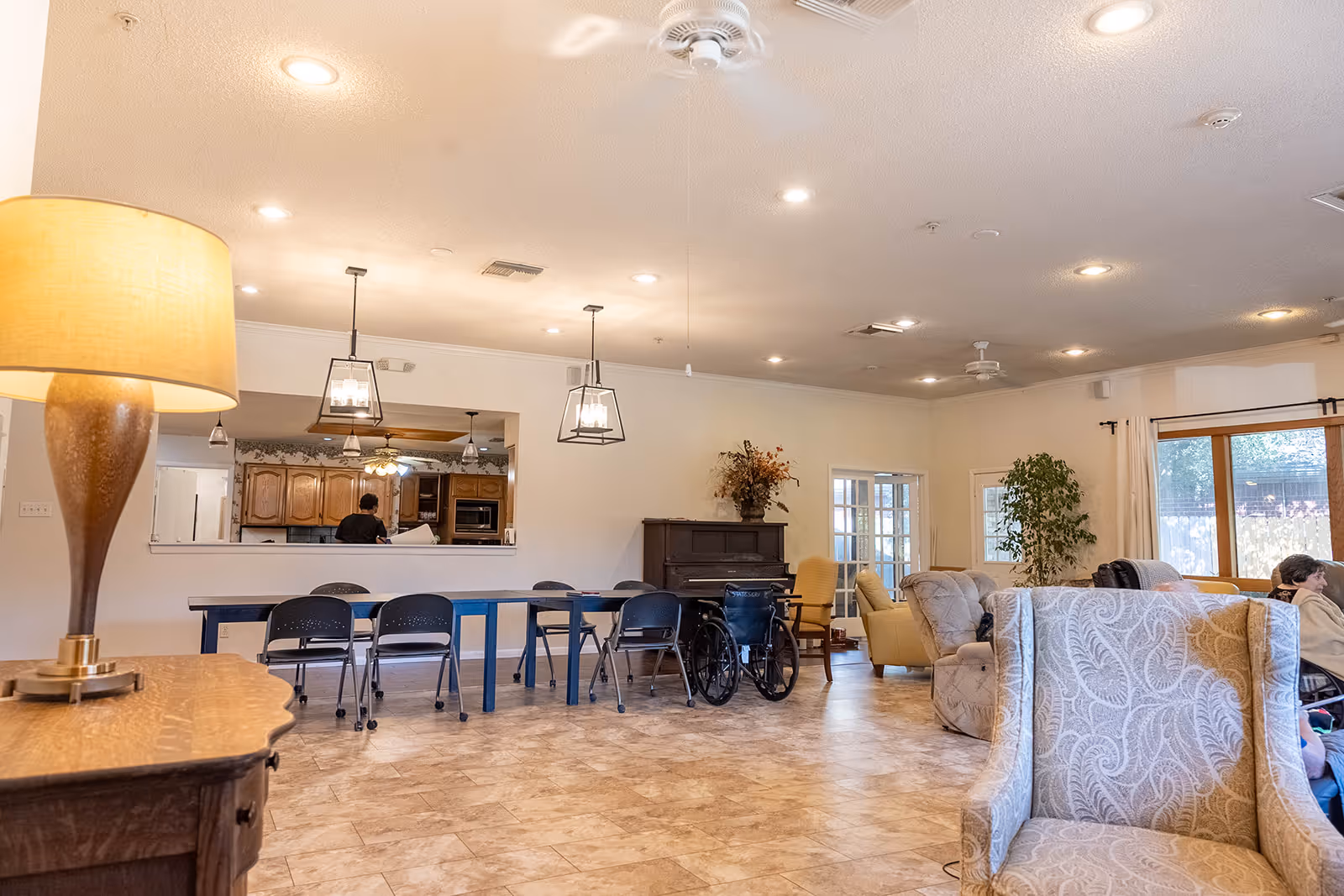 Spacious communal living area with sofas, chairs, dining tables, a wheelchair, and a kitchen pass-through.