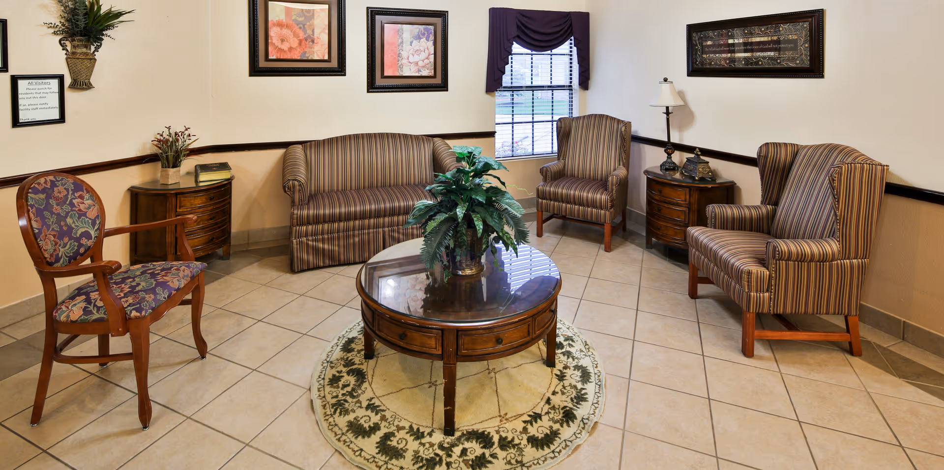 A cozy sitting area in a nursing and rehabilitation center featuring a striped loveseat, two matching striped armchairs, a wooden chair with floral upholstery, a round wooden coffee table with a plant centerpiece, two small wooden side tables with decorative items, framed floral artwork on the walls, a window with blinds and a dark valance, and tiled flooring with a round patterned rug under the coffee table.