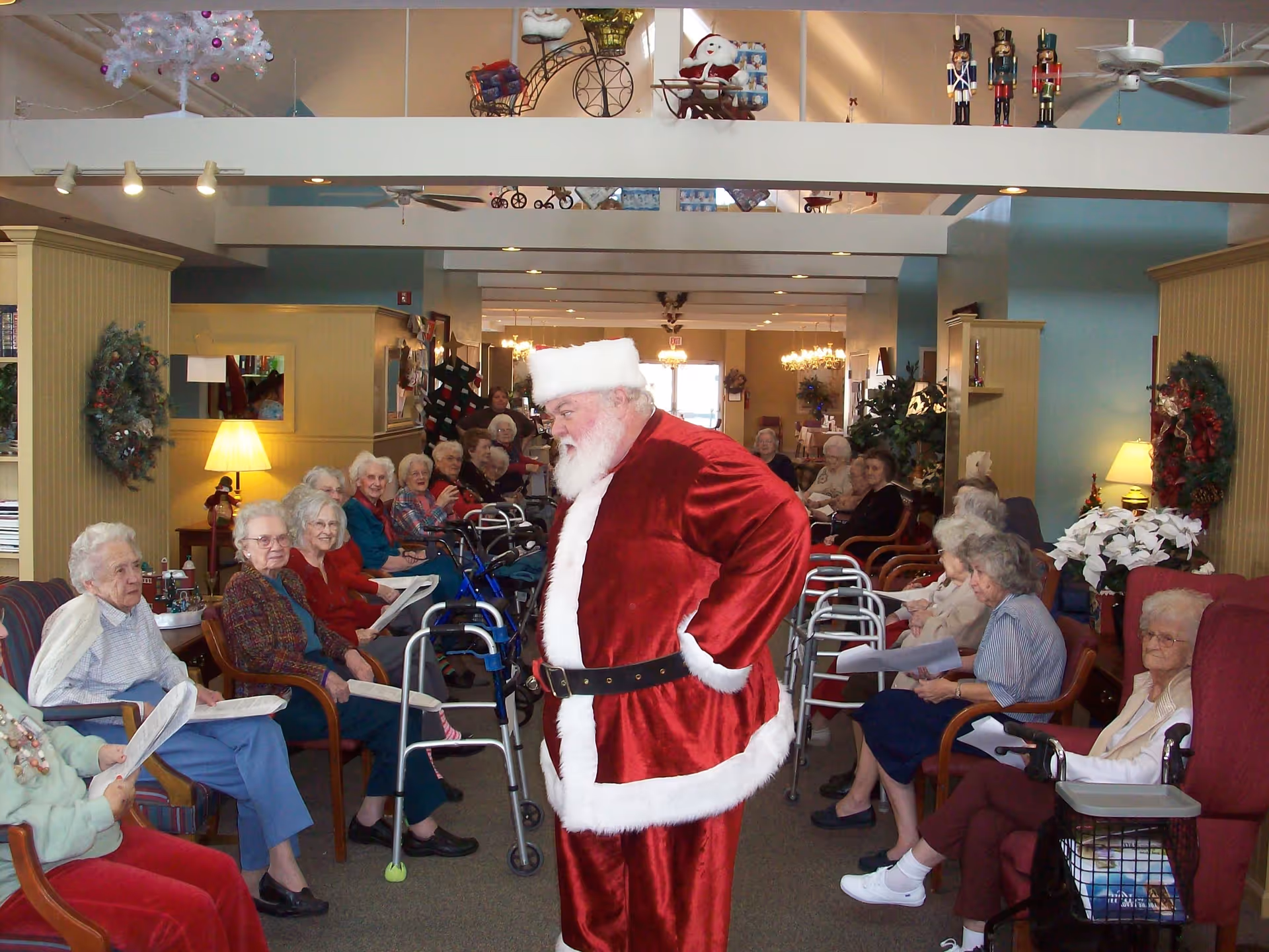 Santa Claus in a red suit stands in a senior living facility common room while elderly residents sit around him.