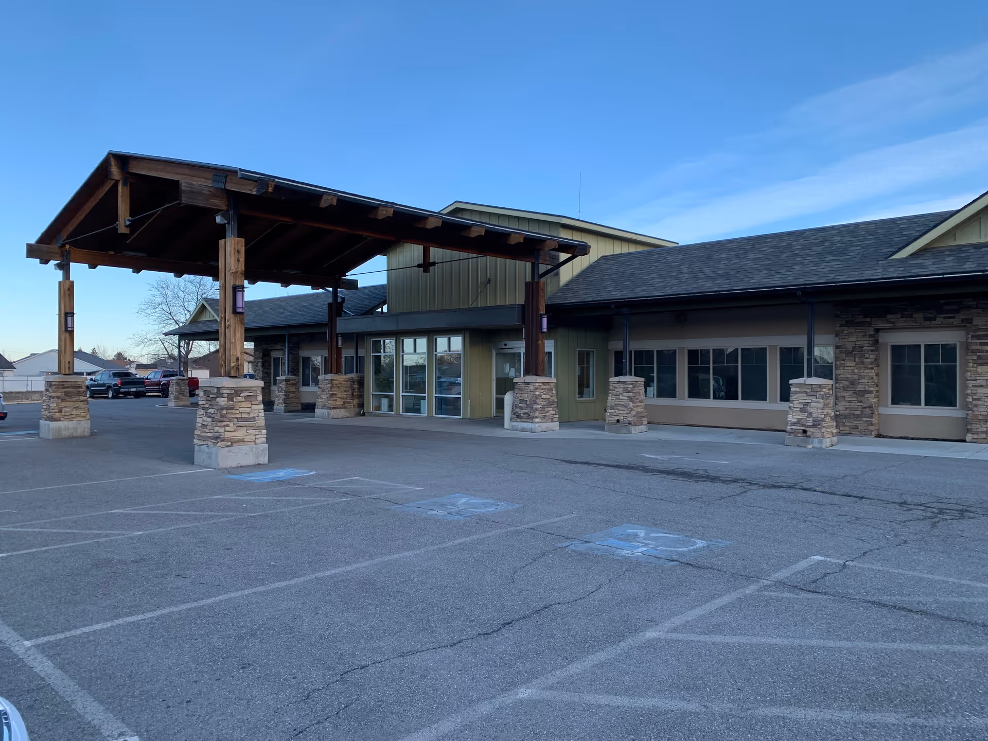Exterior view of Lomond Peak Nursing and Rehabilitation facility showing the entrance with a large wooden canopy supported by stone pillars, adjacent to a parking lot with several handicap parking spaces marked on the asphalt.