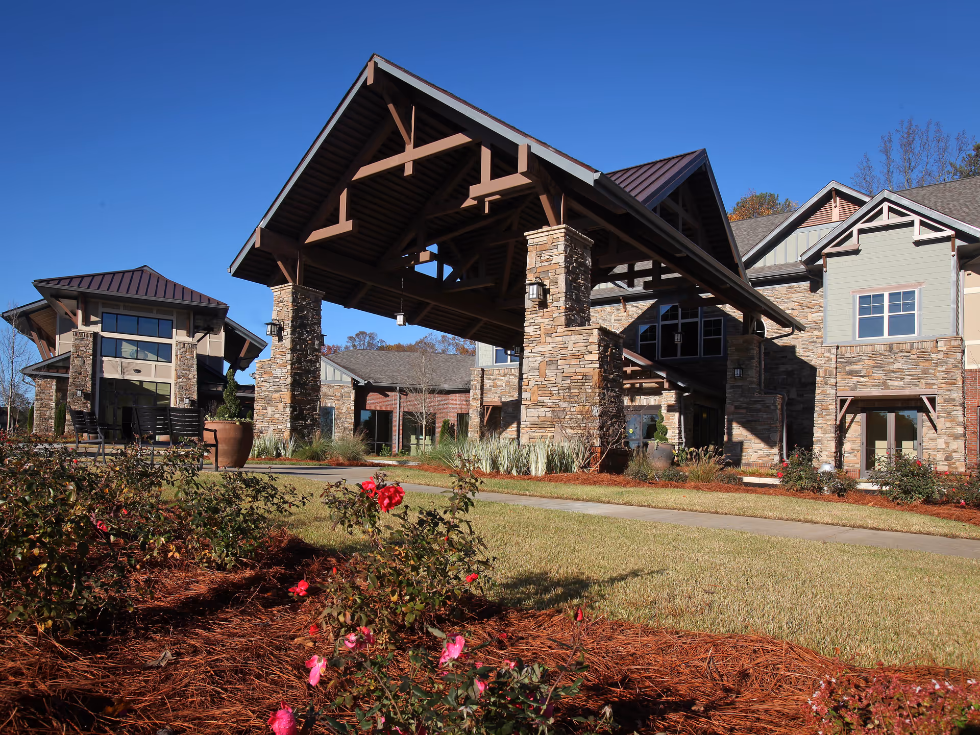 Exterior view of a senior living facility with a large covered entrance supported by stone pillars, surrounded by landscaped gardens with blooming flowers and a clear blue sky.