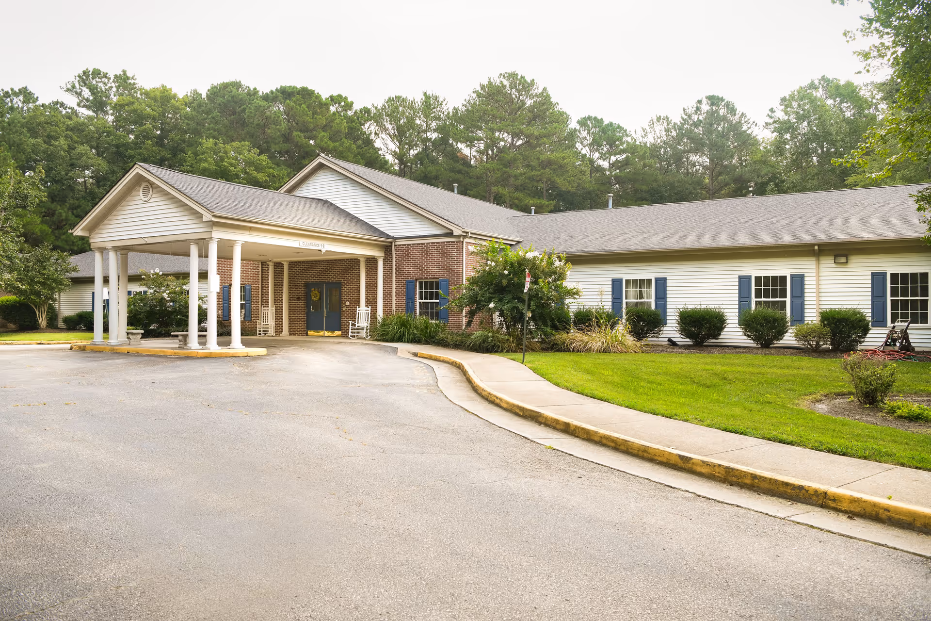 Exterior view of a single-story senior living facility building with a covered entrance supported by white columns, surrounded by green grass, bushes, and trees.