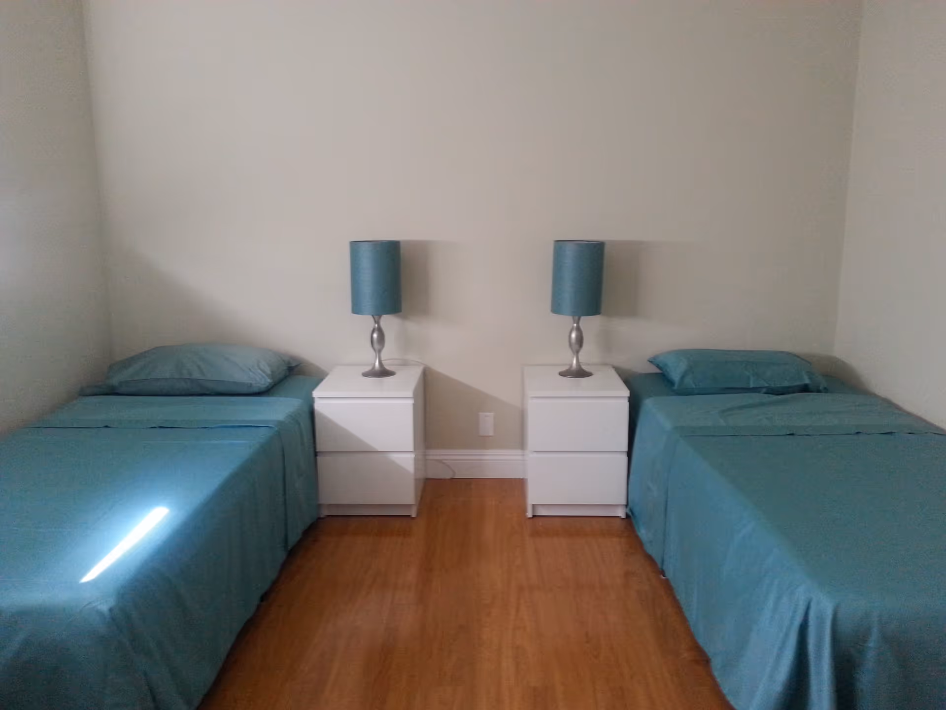 A simple bedroom with two single beds placed parallel to each other, each with teal bedding and pillows. Between the beds are two white nightstands, each holding a silver lamp with a teal lampshade. The room has light-colored walls and wooden flooring.