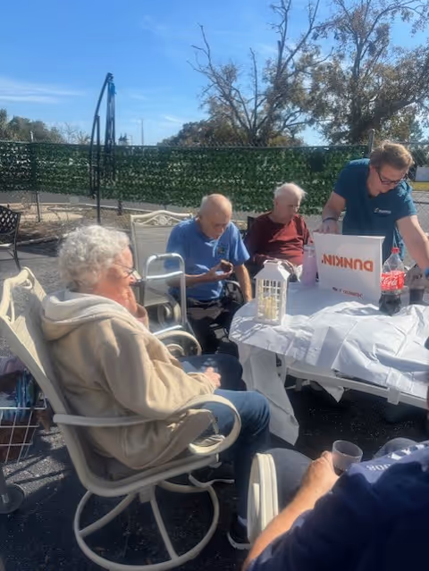 A group of elderly people sitting outdoors around a table covered with a white tablecloth. One person is standing and appears to be serving or organizing items on the table, which includes a Dunkin' box and a bottle of Coca-Cola. The setting is sunny with a green hedge and trees in the background.