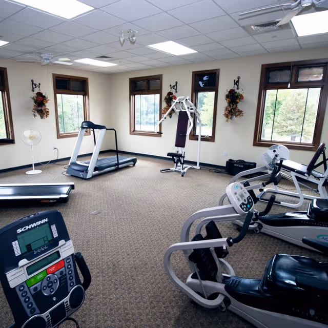 A fitness room with exercise equipment including a treadmill, stationary bike, and weight machine. The room has beige walls, carpeted floor, several windows with wooden frames, and decorative wall hangings with flowers. There is also a standing fan near one of the windows.