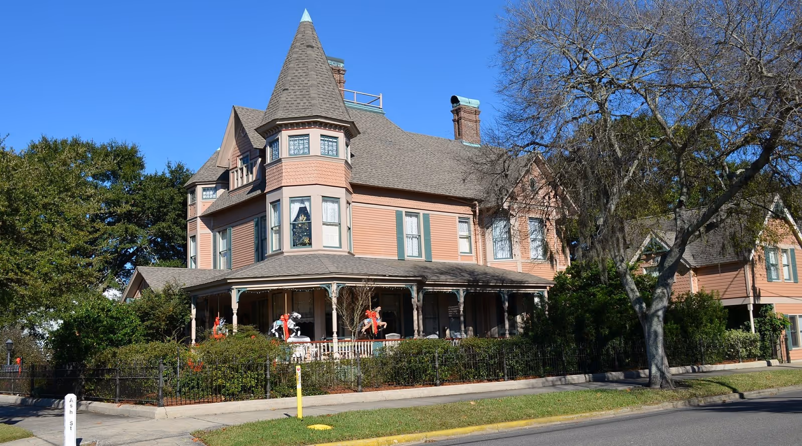Victorian-style pink house with a turret and wraparound porch surrounded by trees and a fence under a clear blue sky.