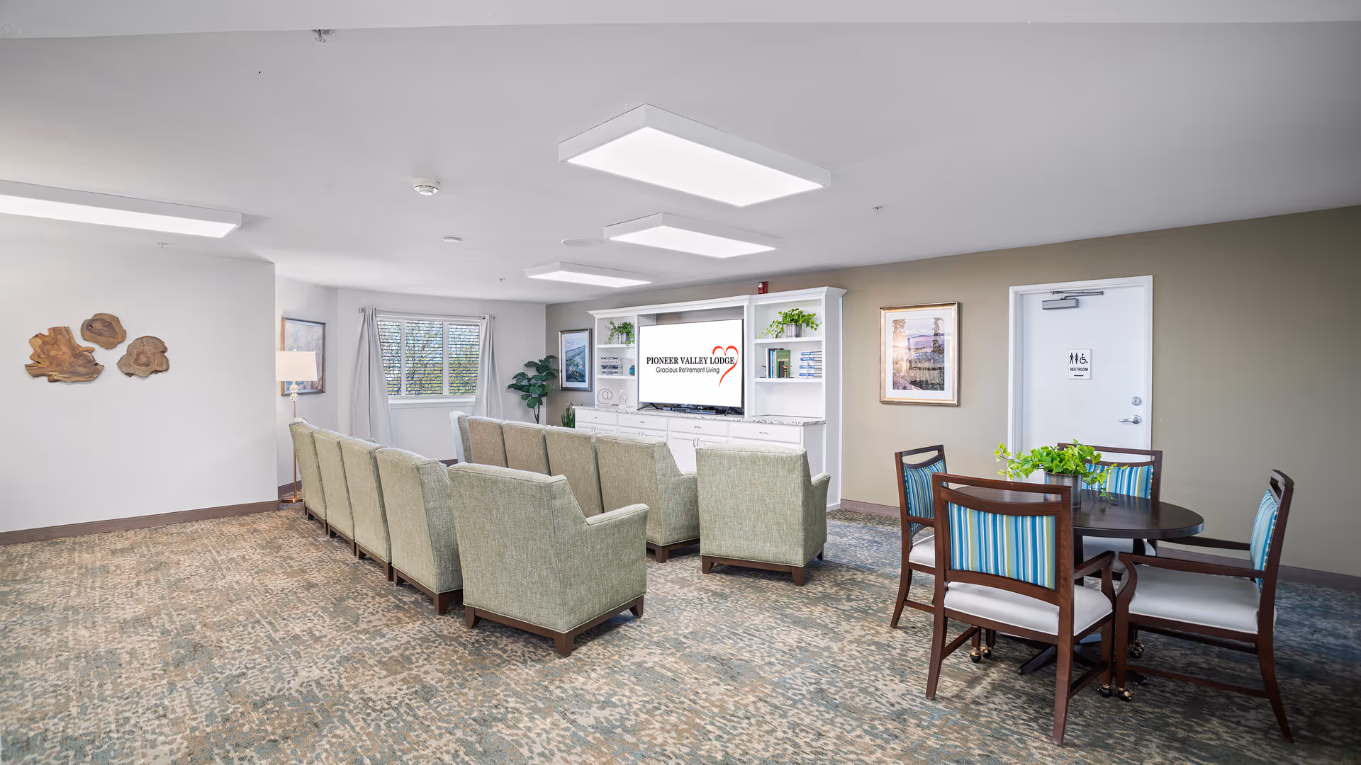 A bright and spacious common living area in a senior living facility with two rows of green upholstered armchairs facing a large white entertainment center with a TV displaying the Pioneer Valley Lodge logo. To the right, there is a round table with four wooden chairs featuring blue striped cushions and a green plant centerpiece. The room has patterned carpet, neutral walls, ceiling lights, framed artwork, and a window with white curtains letting in natural light. A door labeled restroom is visible on the right wall.