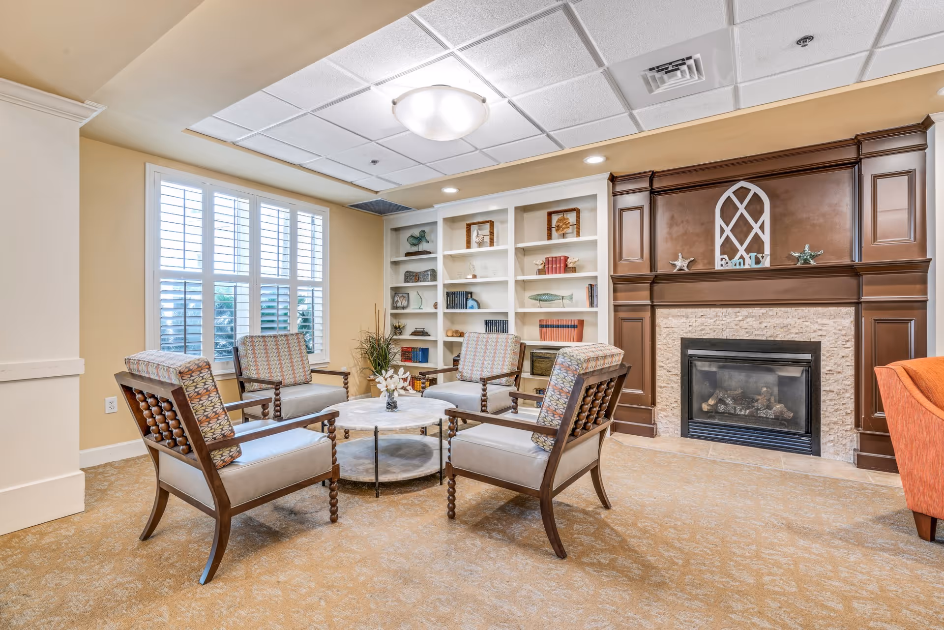 A cozy living room area with four wooden armchairs arranged around a round marble coffee table. Behind the chairs is a large window with white shutters, and to the right is a dark wood fireplace with a stone surround. Built-in white shelves filled with decorative items and books flank the fireplace. The room has beige walls and carpeted flooring.