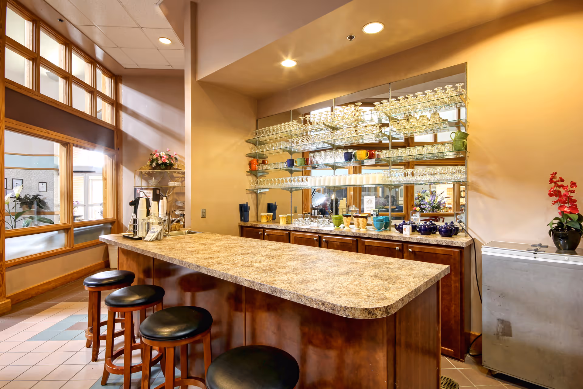 Indoor communal bar area with a long marble-top counter, four stools, shelves of glassware and cabinets, and large windows.