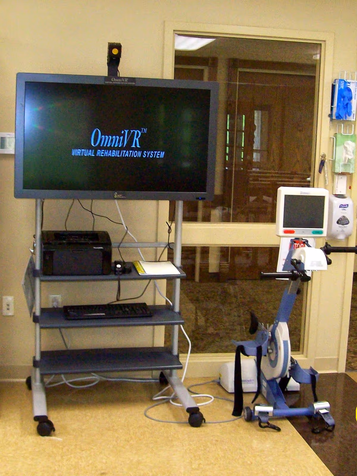 A television on a rolling cart displaying 'OmniVR Virtual Rehabilitation System' next to a stationary exercise bike inside a therapy room.