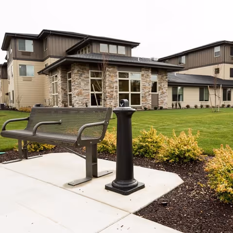 Outdoor scene at Bonaventure of Albany showing a metal bench and a black drinking fountain on a concrete path, with a well-maintained lawn, shrubs, and a modern multi-story building with stone and beige siding in the background.
