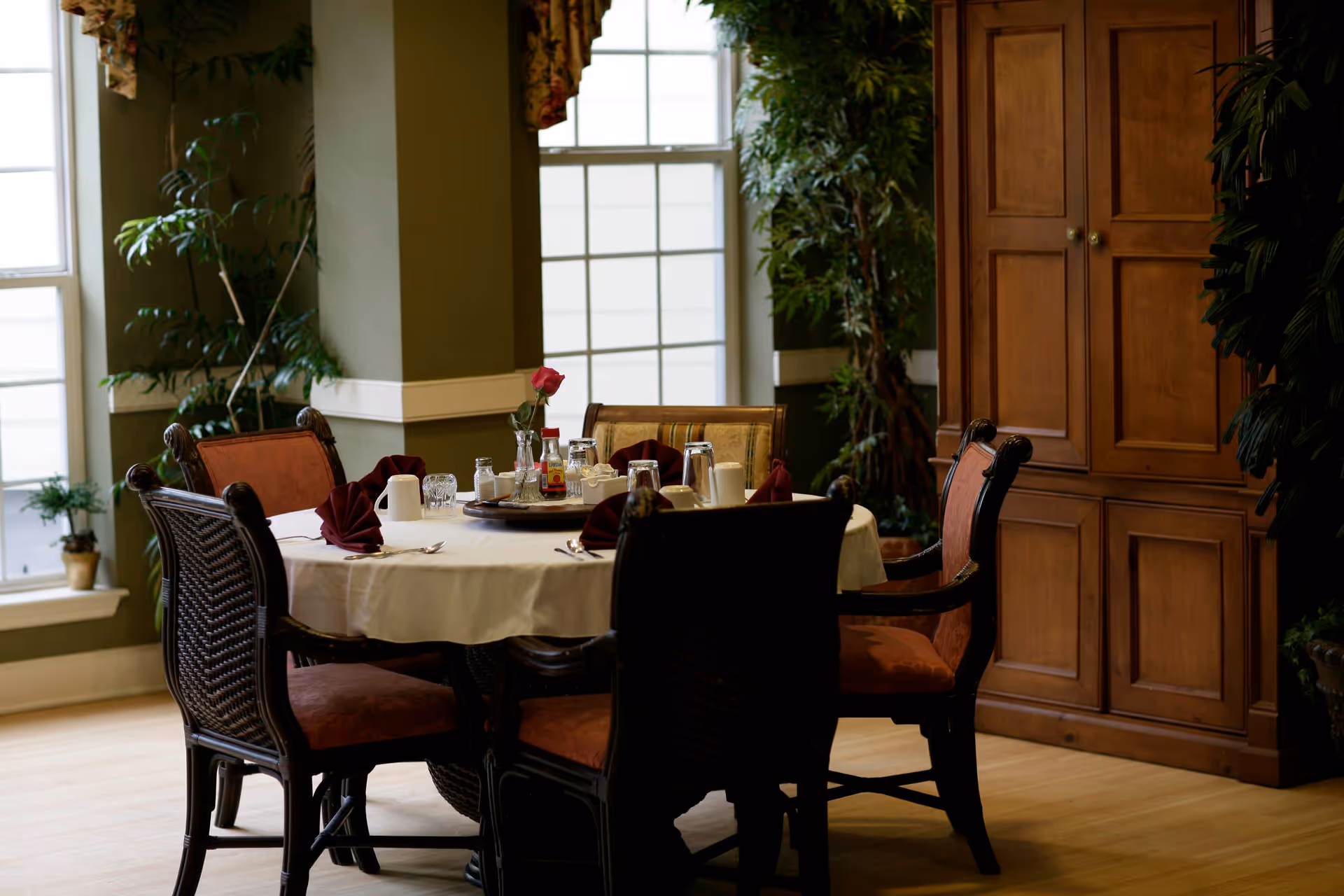 A dining room with a round table set for a meal, featuring a white tablecloth, folded burgundy napkins, glassware, condiments, and a single red rose in a vase. The room has large windows with floral curtains, green walls, wooden flooring, and a wooden cabinet. There are six cushioned chairs around the table and several potted plants in the background.