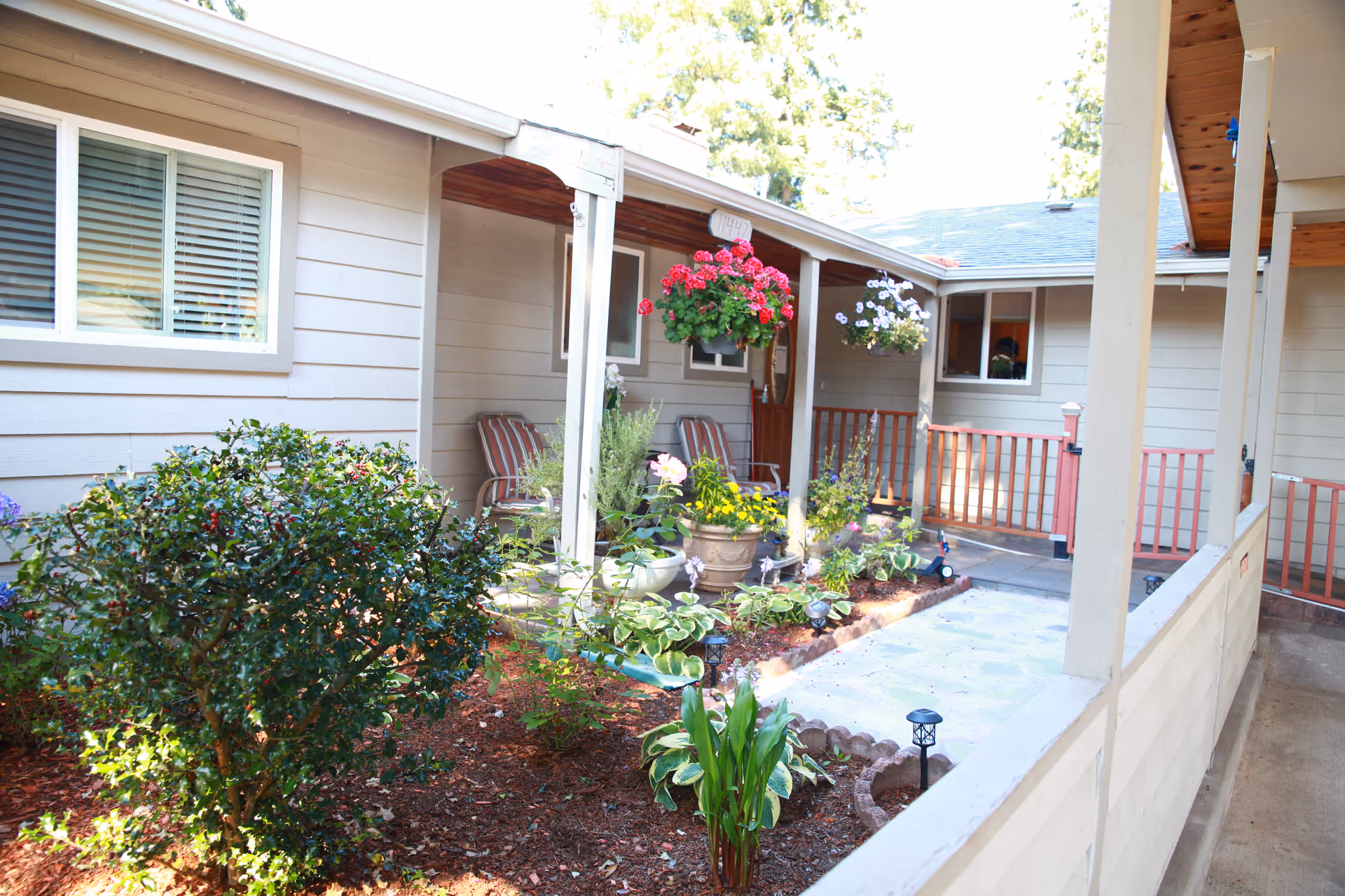 Outdoor courtyard area of a senior living facility with a garden bed containing various plants and flowers. There are hanging flower pots with pink and white flowers, striped chairs along the wall of the building, and a covered walkway with wooden posts and railings.