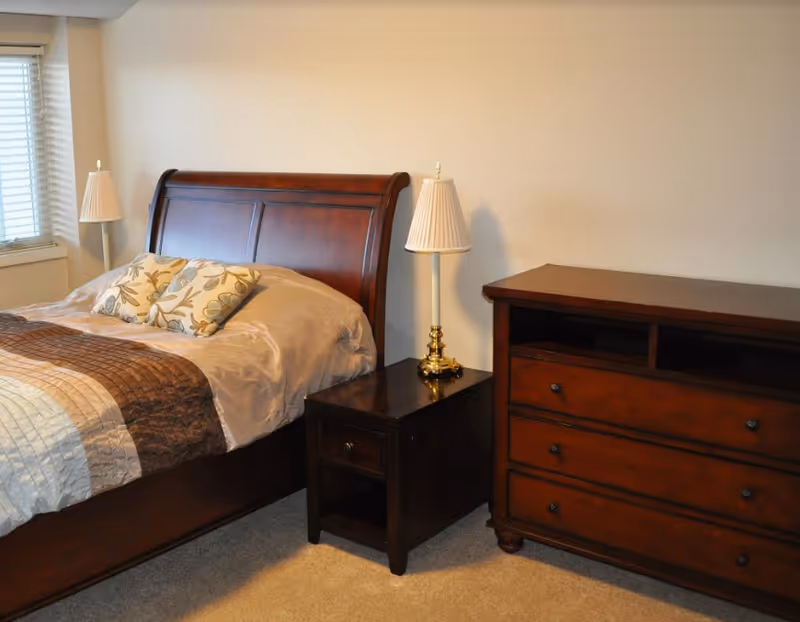 A bedroom with a wooden bed frame featuring a beige and brown quilt, two floral pillows, a wooden nightstand with a brass lamp, and a wooden dresser against a beige wall.