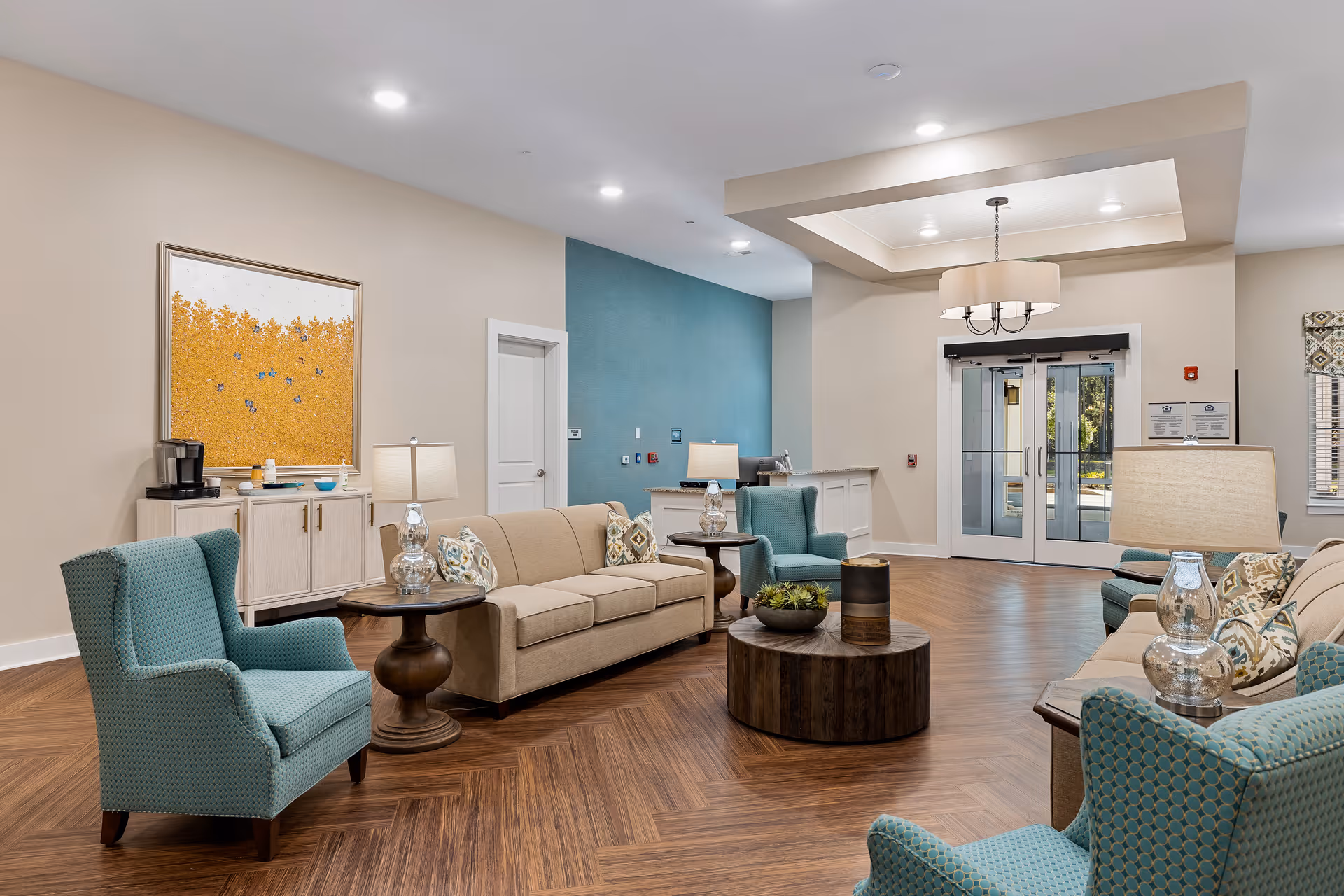 Bright, modern senior living lobby with beige sofas, teal armchairs, round coffee tables, lamps, and glass entrance doors.