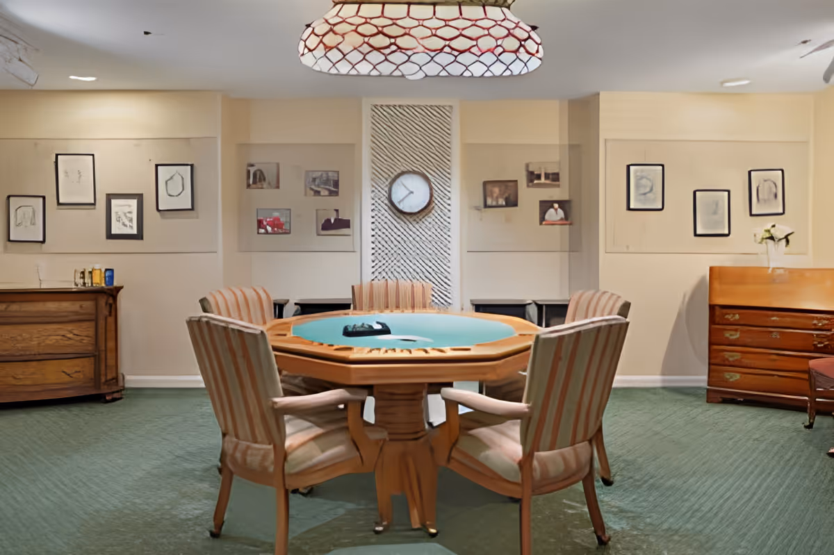 A cozy room with a round wooden table in the center surrounded by six striped upholstered chairs. The table has a green felt top and a tray with some items on it. The room has green carpet flooring, beige walls adorned with framed pictures and a clock in the middle. There are wooden cabinets on either side of the room and a large light fixture hanging from the ceiling.