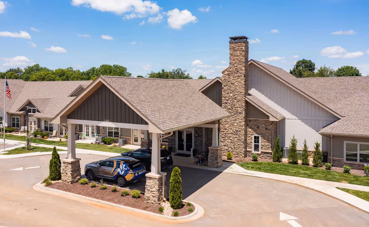 Front entrance of a senior living facility featuring a covered porte-cochere with stone columns, a tall stone chimney, landscaping, and two parked cars.