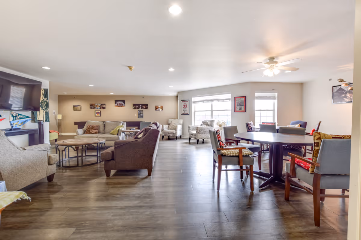 A bright and spacious senior living community lounge area with multiple seating options including sofas, armchairs, and a round table with chairs. The room features large windows allowing natural light, ceiling lights, and a ceiling fan. The walls are decorated with framed pictures and sports memorabilia, and there is a flat-screen TV mounted on the wall.
