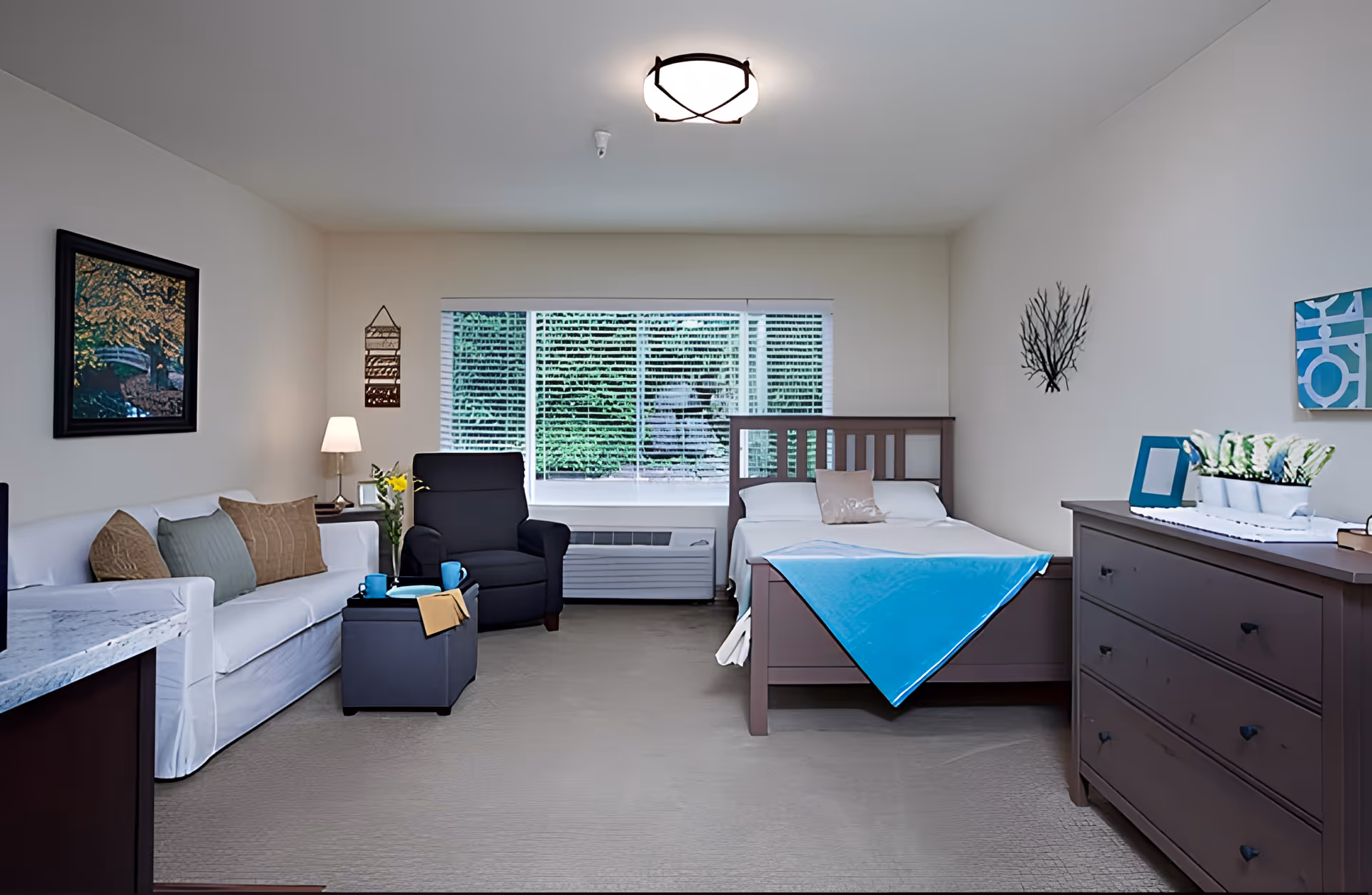 A well-lit senior living bedroom featuring a wooden bed with a blue blanket, a white sofa with cushions, a black armchair, a small ottoman with two blue mugs and a yellow napkin, a wooden dresser with decorative items, a window with blinds showing greenery outside, and wall art including a framed picture and a coral decoration.