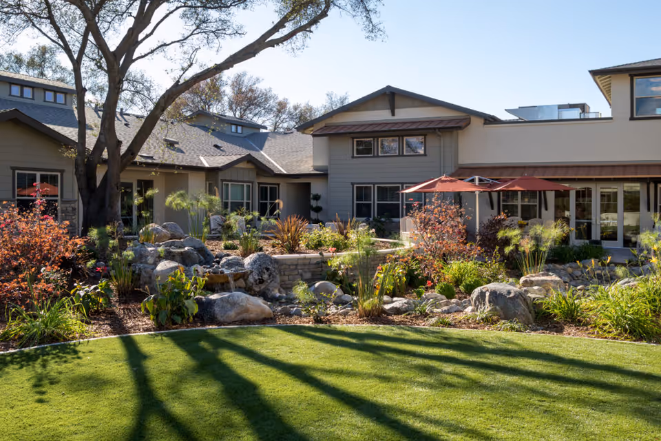 A landscaped garden area with green grass, rocks, shrubs, and small trees in front of a residential-style building with multiple windows and a patio umbrella providing shade.