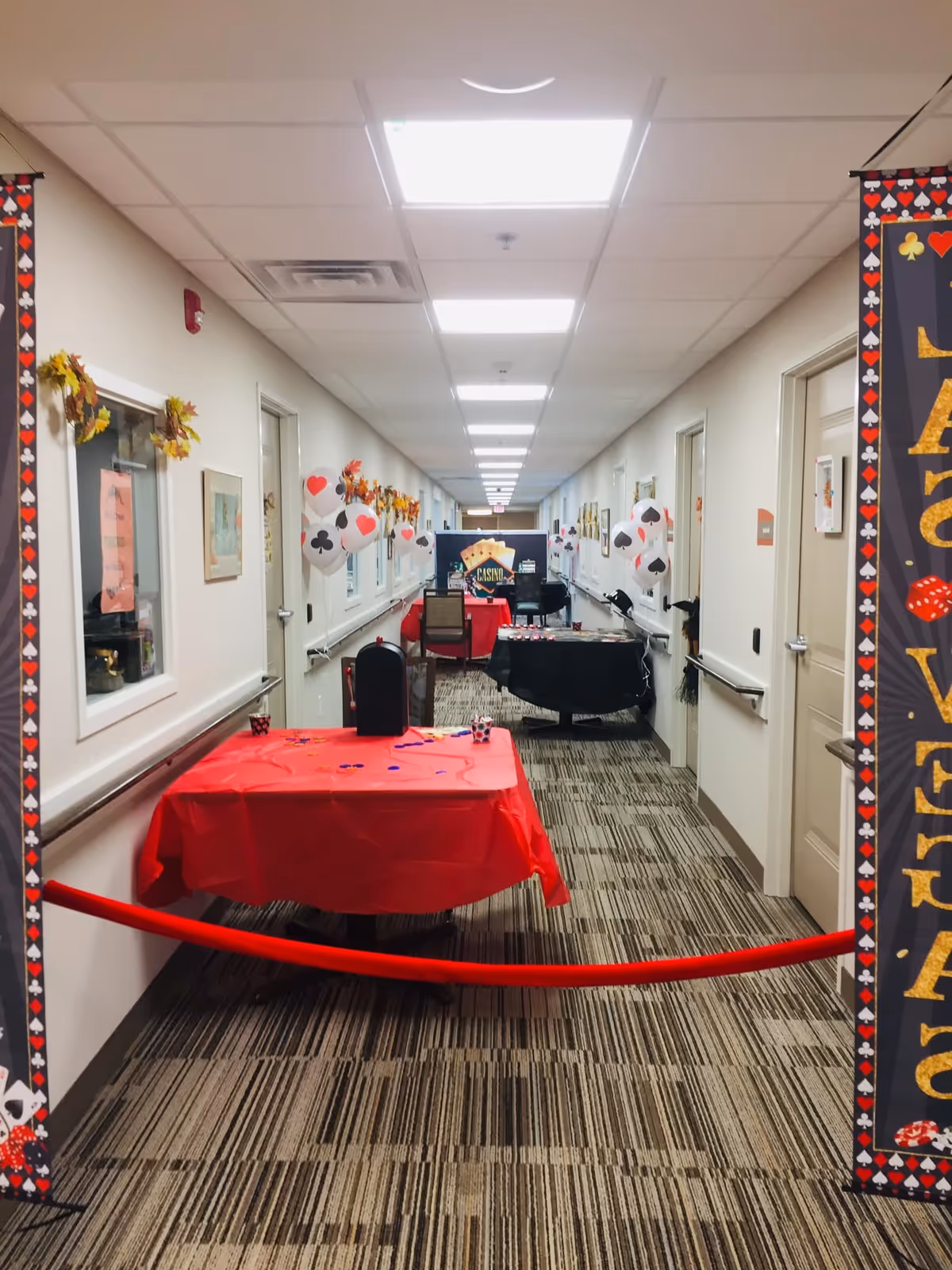 A decorated hallway in an assisted living facility with tables covered in red and black tablecloths set up for a casino-themed event. The walls are adorned with playing card suit balloons and autumn wreaths. A red velvet rope blocks the entrance to the hallway.