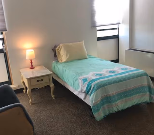 A simple bedroom with a single bed covered with a turquoise and white patterned blanket and a beige pillow. Next to the bed is a small vintage-style nightstand with a lit lamp on it. The room has carpeted flooring and two windows with blinds partially drawn.
