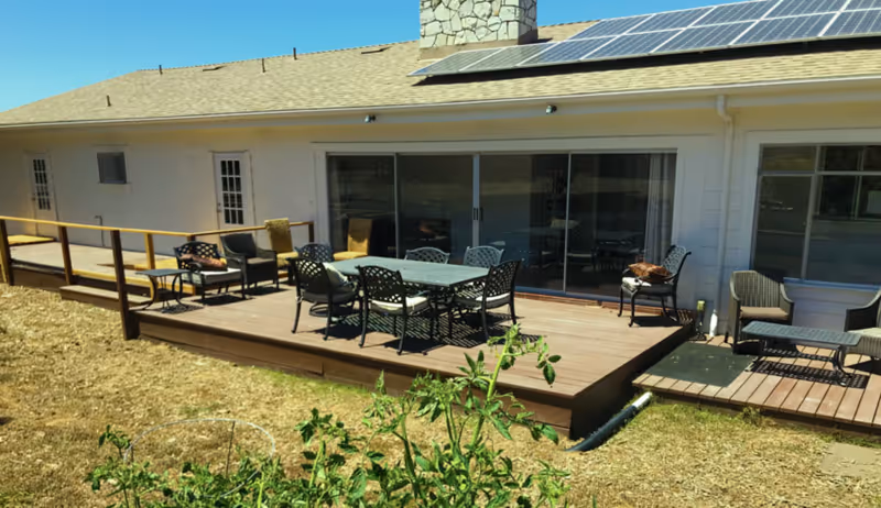Outdoor wooden deck attached to a single-story building with solar panels on the roof. The deck has several chairs and tables arranged for seating. There is a small garden area with plants in the foreground and a clear blue sky above.