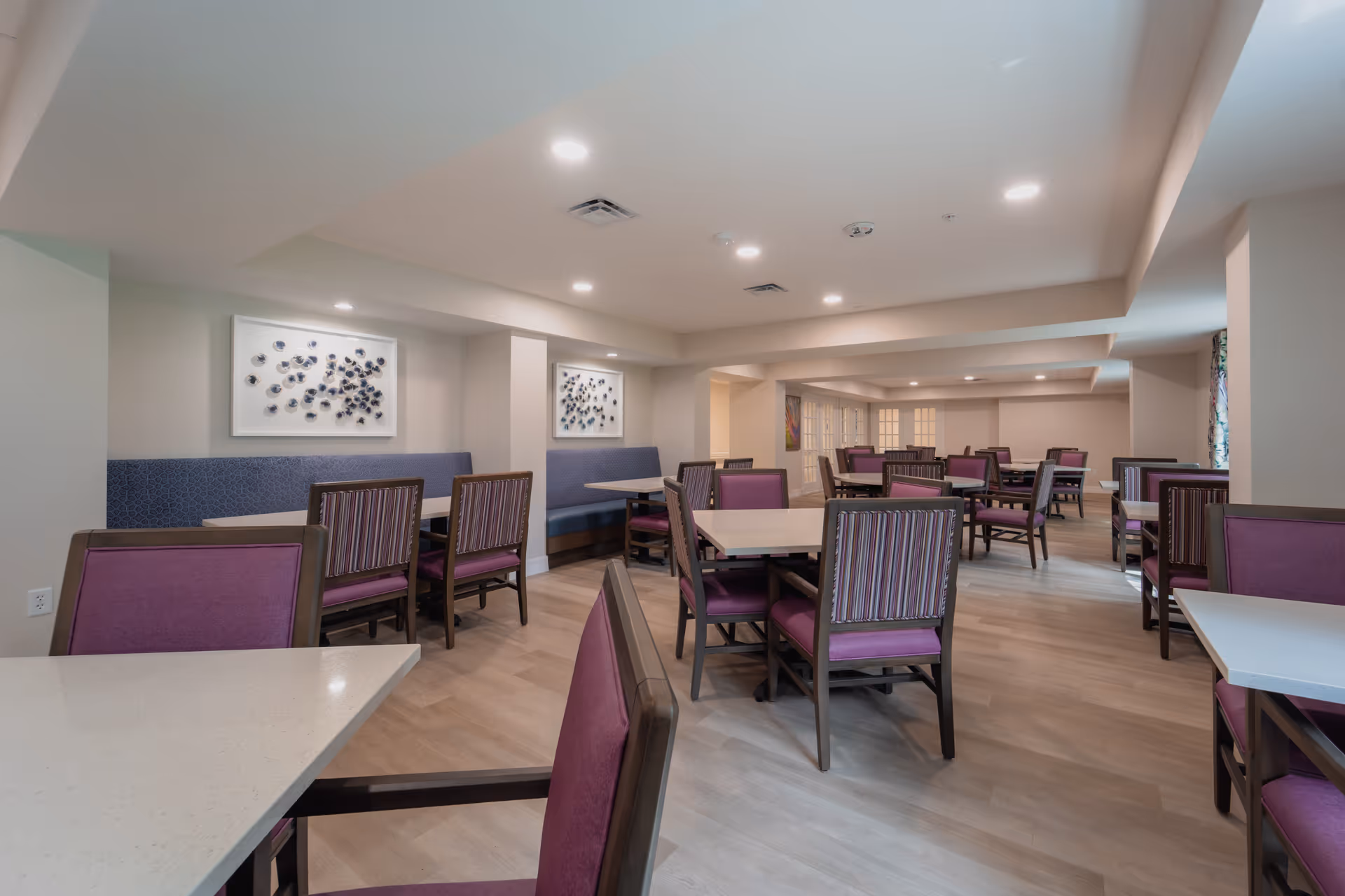 A spacious dining room with multiple tables and chairs featuring purple upholstery and striped backs. The room has light-colored walls, recessed ceiling lights, and decorative wall art. The floor is a light wood finish, and there are booth-style seating areas along the walls.
