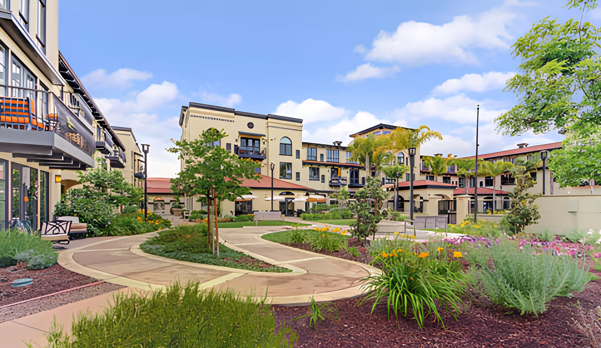 Outdoor garden area with winding paved pathways, various plants, flowers, and small trees surrounded by multi-story residential buildings under a partly cloudy sky.