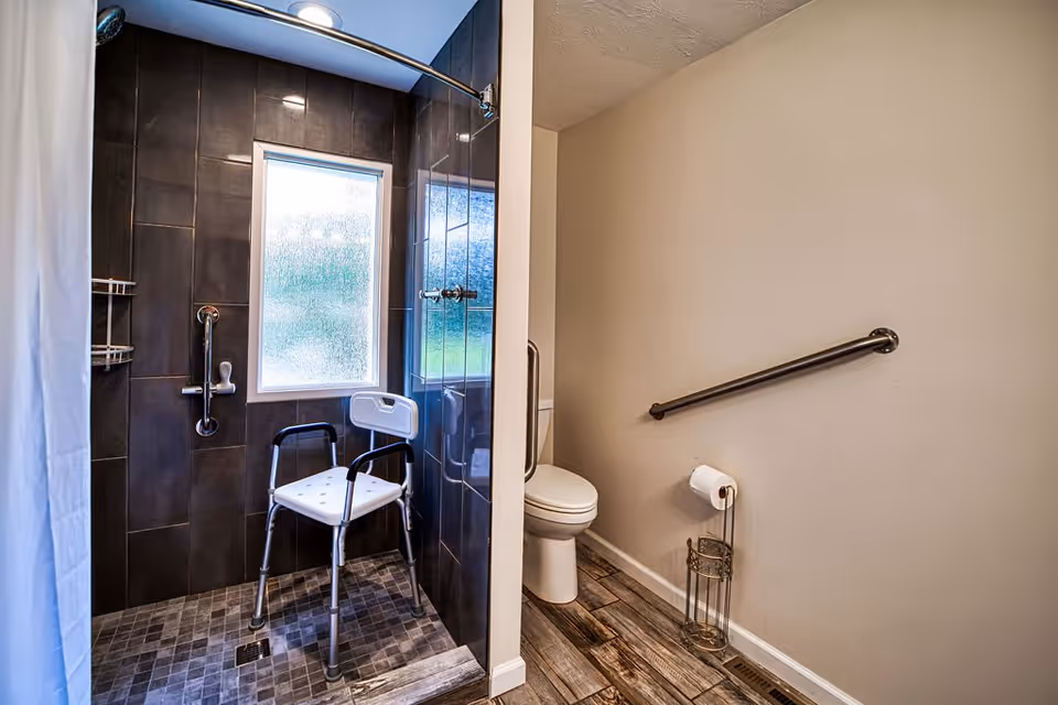 Accessible bathroom with a walk-in shower featuring dark tiles, a white shower chair, grab bars, and a frosted window. Adjacent to the shower is a toilet with a grab bar on the wall and a toilet paper holder on the floor. The floor has wood-like tiles and the walls are painted beige.