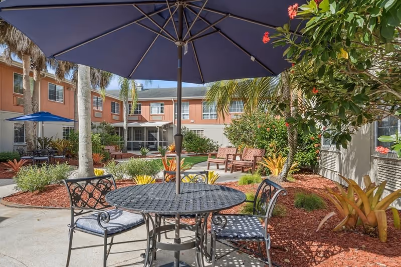 Outdoor courtyard area with a round metal table and four chairs under a large blue umbrella. The courtyard is surrounded by a two-story building with windows, palm trees, various plants, and additional seating areas with benches and tables under umbrellas.