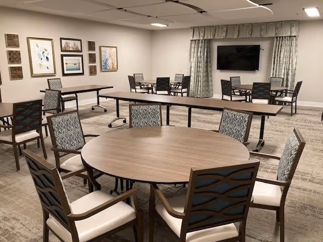 A spacious room with multiple tables and chairs arranged for group seating. The room features a round table with six cushioned chairs in the foreground, several rectangular tables with chairs in the background, framed artwork on the walls, a wall-mounted flat-screen TV, and patterned curtains covering a window.