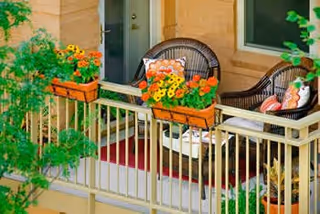 A small balcony with two wicker chairs, colorful flower boxes on the railing, and a door and window leading inside.