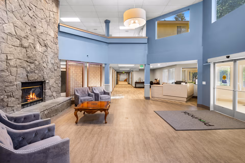 Spacious senior living community lobby with a stone fireplace on the left, three gray upholstered chairs around a wooden coffee table, a reception desk on the right, and large windows letting in natural light.