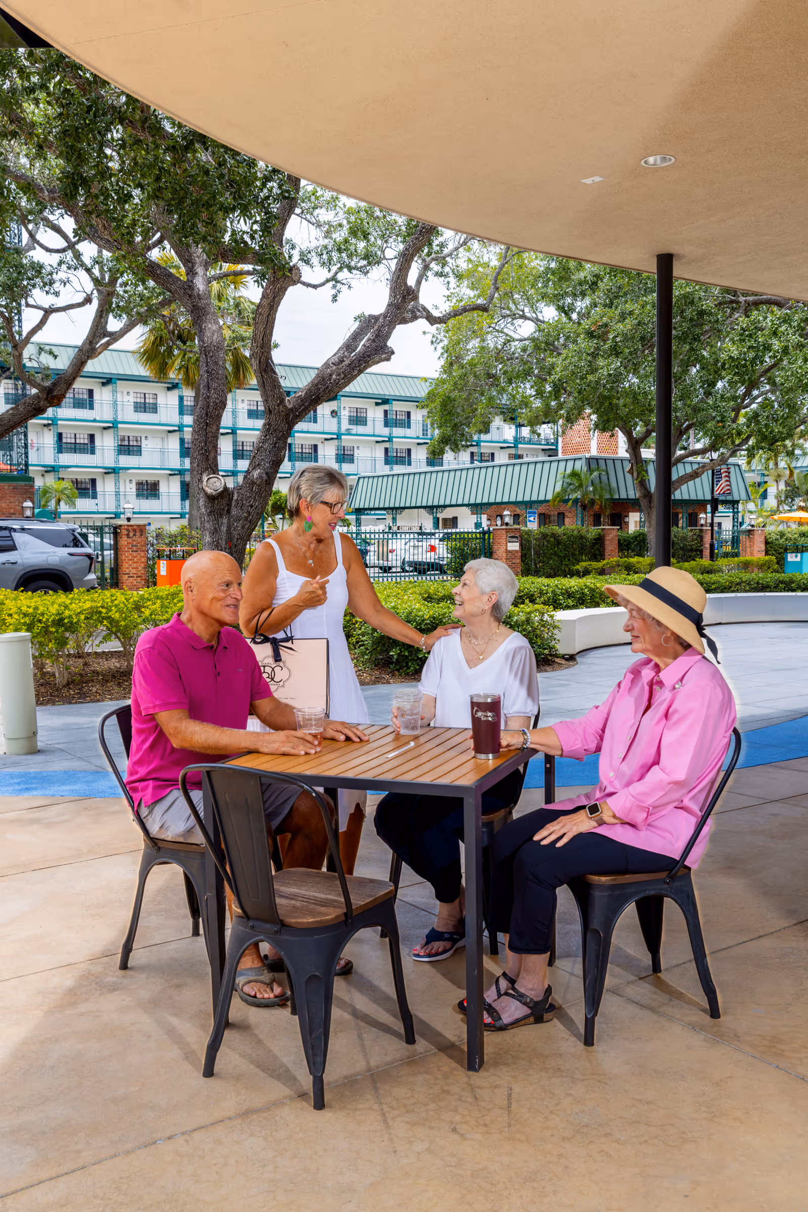 Four elderly people socializing outdoors at a senior living facility. Three are seated around a wooden table with metal chairs, holding drinks, while one woman stands and interacts with the group. The setting includes trees, bushes, and a multi-story building in the background.