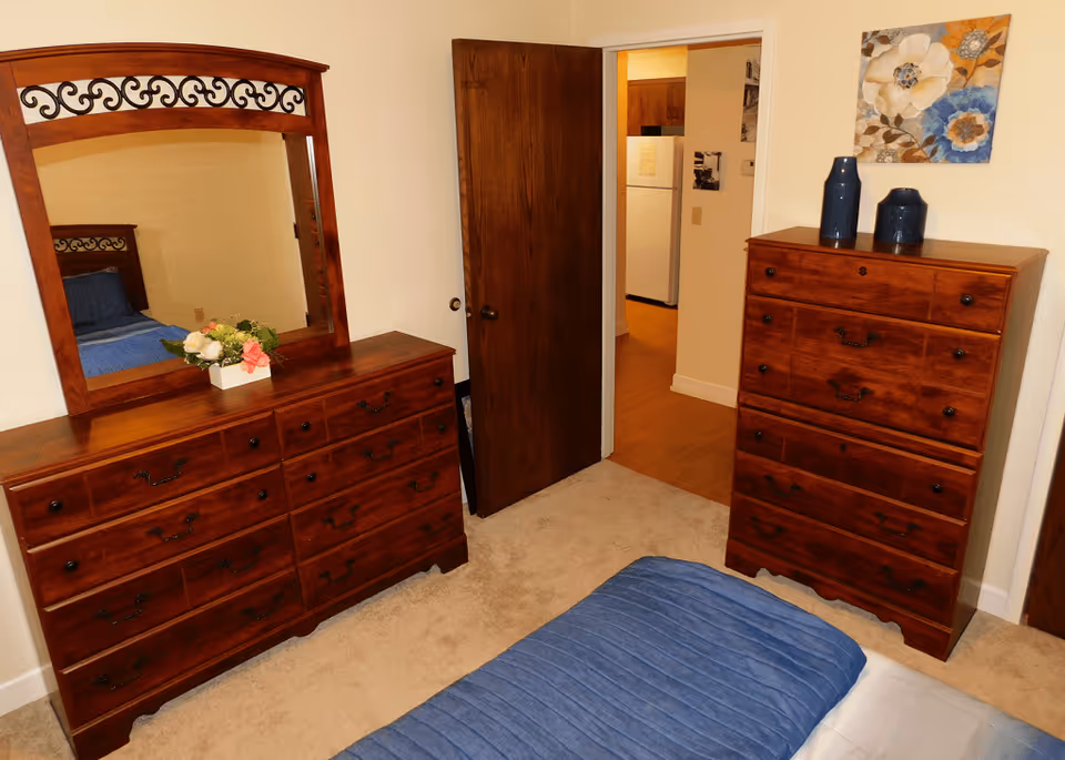 A bedroom with wooden furniture including a dresser with a large mirror and a chest of drawers. The bed has a blue quilted blanket. There is a door open leading to a kitchen area with a white refrigerator. A floral painting and two blue vases are on top of the chest of drawers.