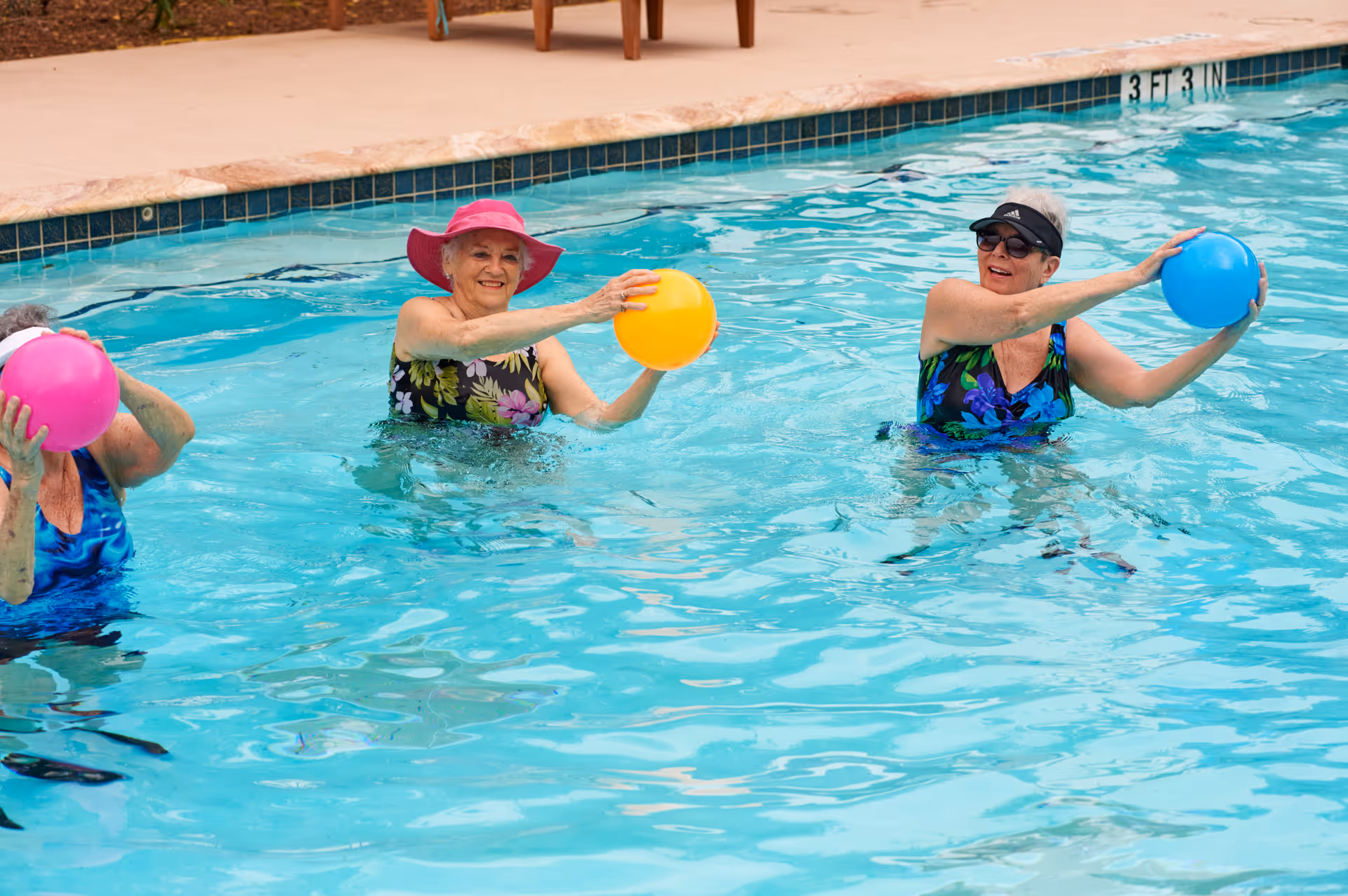 Three elderly women in a swimming pool participating in a water exercise activity, each holding a colorful ball. One woman wears a pink sunhat and floral swimsuit, another wears a black visor and sunglasses with a floral swimsuit, and the third woman is partially visible holding a pink ball.