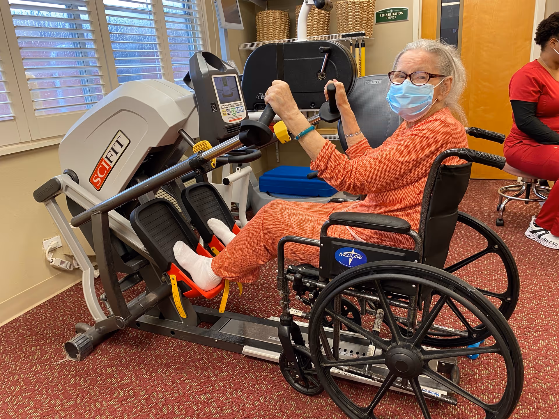 An elderly woman wearing glasses and a face mask is seated in a wheelchair while using a leg exercise machine in a rehabilitation office. She is dressed in an orange outfit and white socks. In the background, a person in red scrubs is seated on a stool near a door, and there are windows with blinds and baskets on shelves.